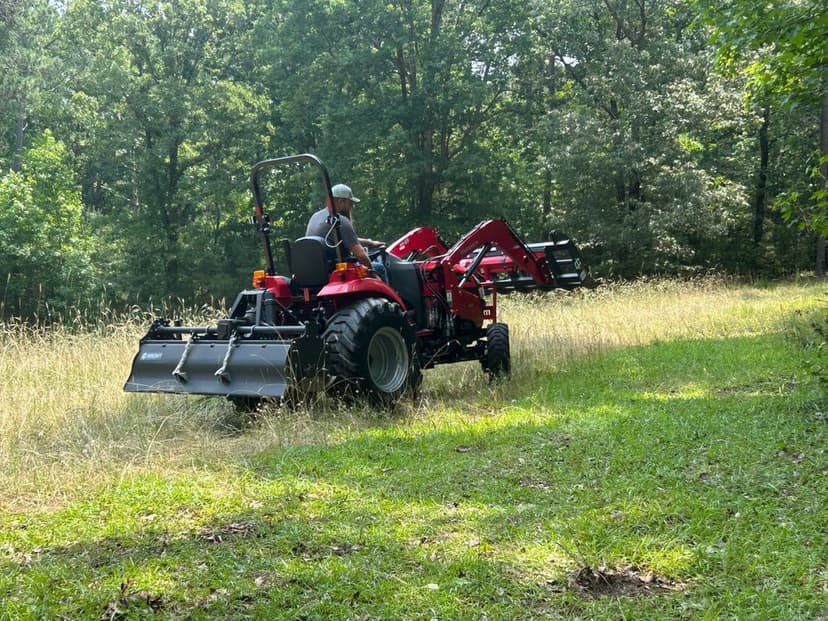 Red tractor mowing tall grass in a forested area, enhancing landscaping.